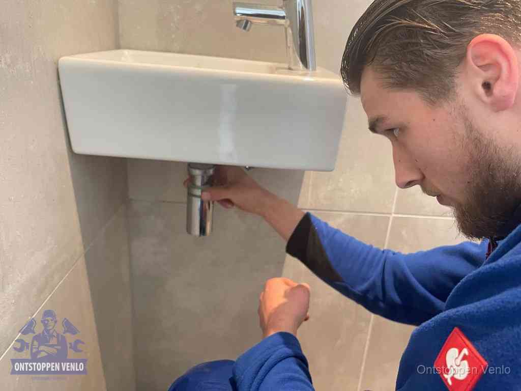 Een monteur draait met de hand de zwanenhalssifon onder een fonteinwasbak in de toiletruimte los om de bron van rioollucht in Venlo te vinden