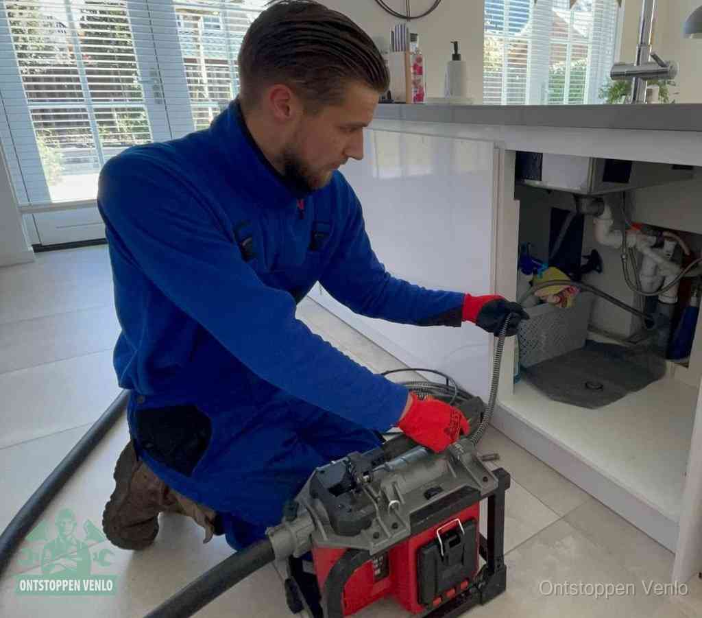 Een monteur met rode handschoenen knielt in een keuken en voert een elektrische ontstoppingsveer in tijdens het professioneel afvoer ontstoppen in Venlo