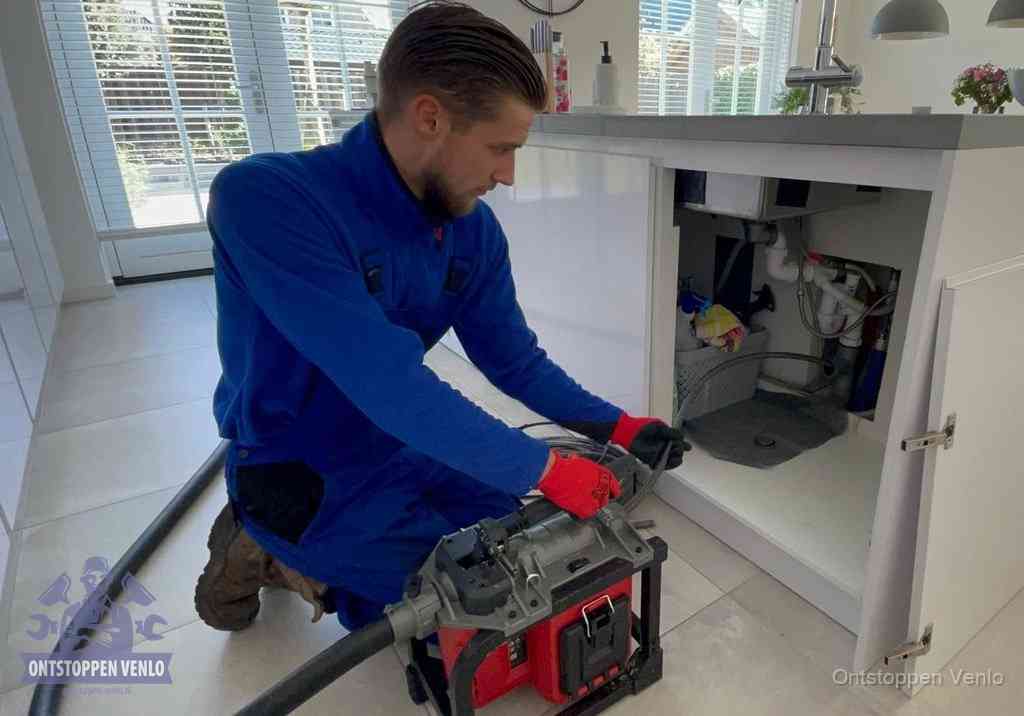 Een monteur voert een professionele elektrische ontstoppingsmachine in de afvoer onder de spoelbak tijdens het gootsteen ontstoppen in Venlo